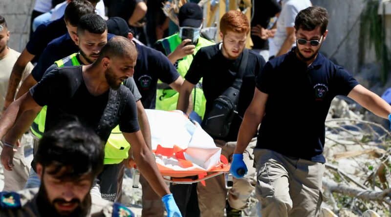 Rescue workers in uniforms and gloves carry a person on a stretcher through rubble at a disaster site.