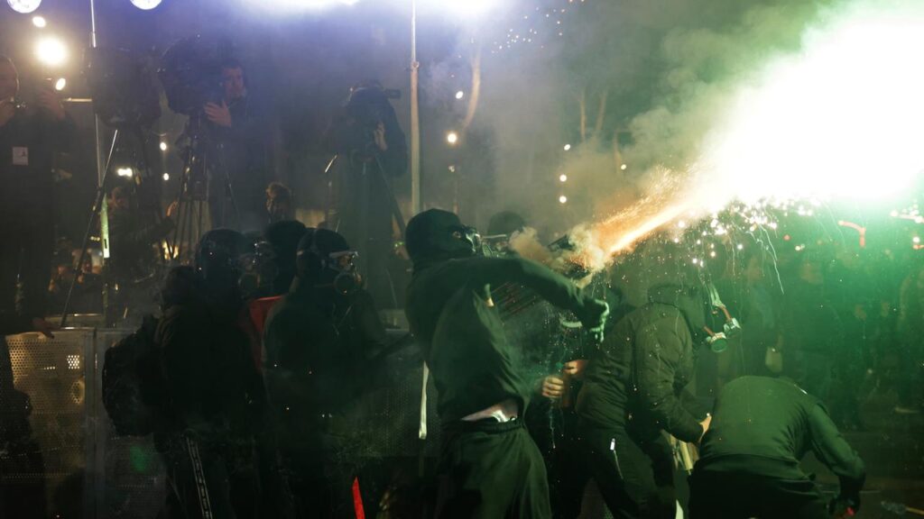 Nighttime scene of masked participants and photographers as a person emits a bright spark toward the crowd, creating smoke and light.