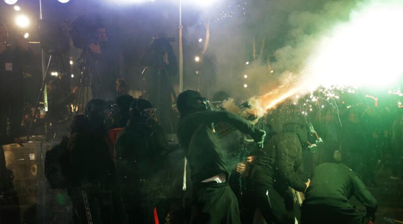 Nighttime scene of masked participants and photographers as a person emits a bright spark toward the crowd, creating smoke and light.