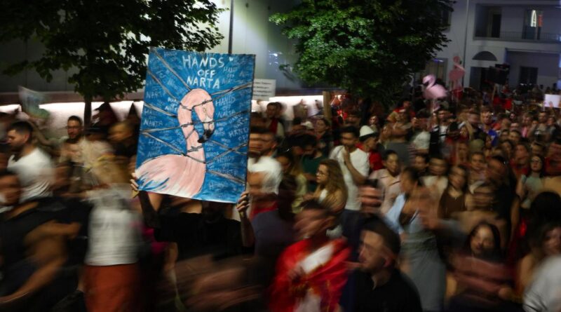 Crowd of protesters marching at night, one person holds a large blue sign reading 'Hands off Narta.'