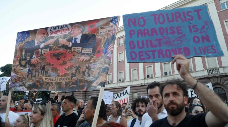 Protest scene with a blue sign reading 'NO TOURIST PARADISE IS BUILT ON DESTROYED LIVES' held above a crowd.
