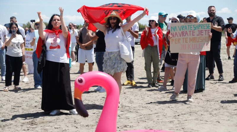 Diverse protesters on a sandy beach hold signs, with a large pink inflatable flamingo in the foreground.