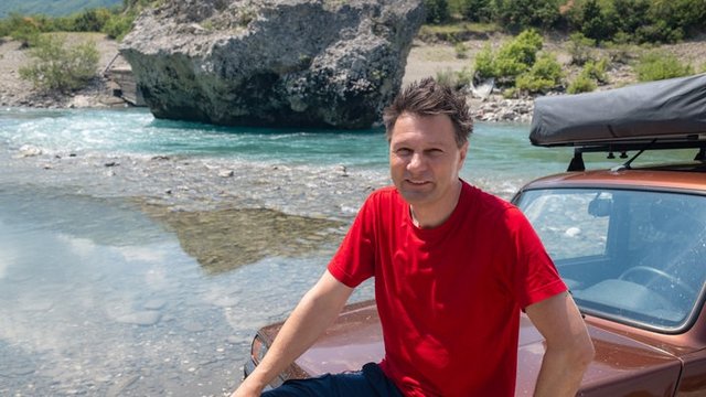 Man in a red T-shirt sits on a brown SUV with a roof rack, by a clear turquoise river and rocky shoreline.