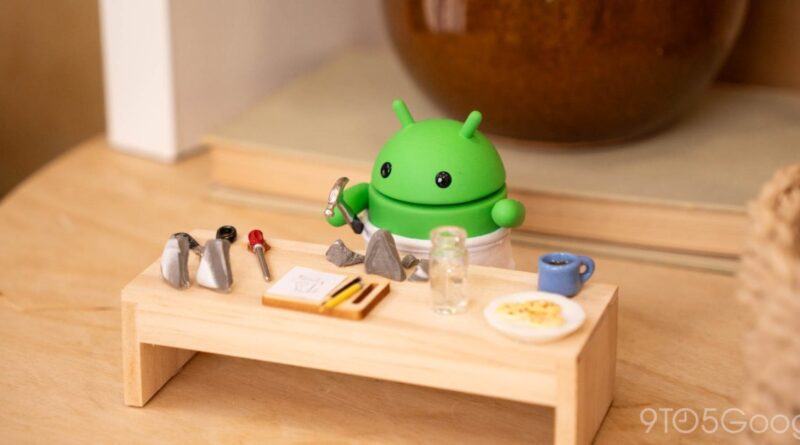 Green Android toy in a white apron beside a tiny wooden workbench with small rocks, tools, and a notebook scattered on the surface.