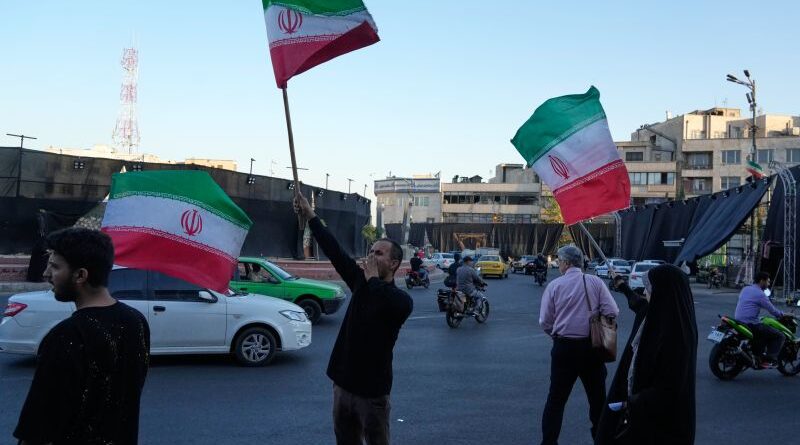 Man in black raises a large Iranian flag on a busy urban street, with cars and motorcycles around him and onlookers nearby.