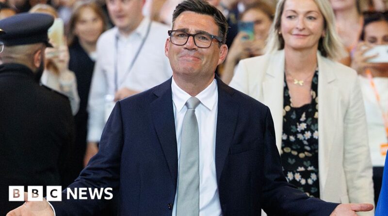 Man in a dark suit and light gray tie, smiling with arms outstretched at a public event; BBC News watermark in bottom left corner.