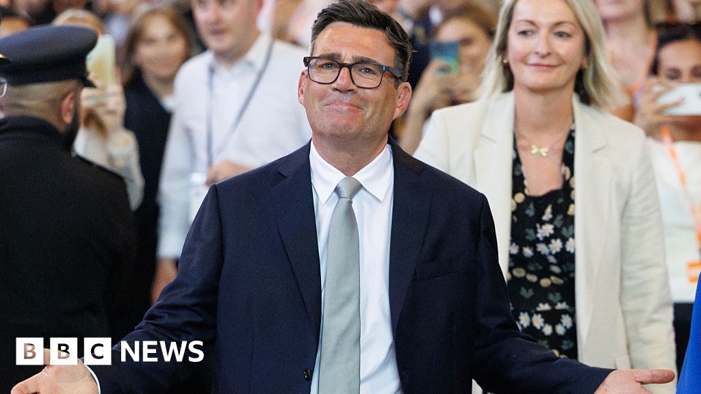 Man in a dark suit and light gray tie, smiling with arms outstretched at a public event; BBC News watermark in bottom left corner.
