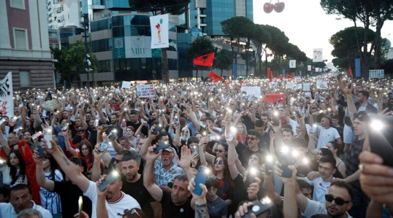 A large outdoor protest with hundreds of people raising illuminated phones, banners and red flags along a city street at dusk.",