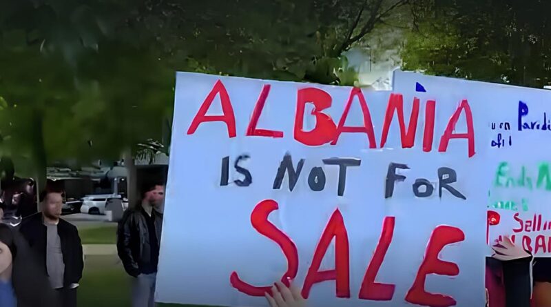 Light blue protest banner reads 'ALBANIA IS NOT FOR SALE' in red and black letters, with people standing in a park-like setting behind it.