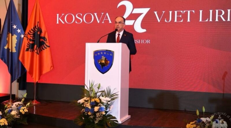 Man in a suit delivering a speech at a white podium bearing a blue shield crest, on a red stage with flags in the background.