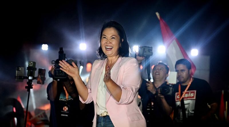 Woman in a pink blazer smiles and claps on a stage as photographers film the moment at a night rally, flag visible in the background.