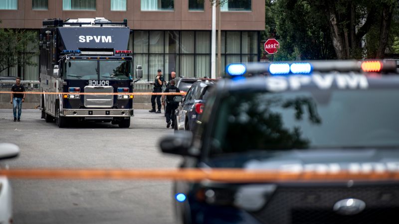 Police and emergency vehicles at a cordoned-off scene with flashing lights and orange tape in front of a building.