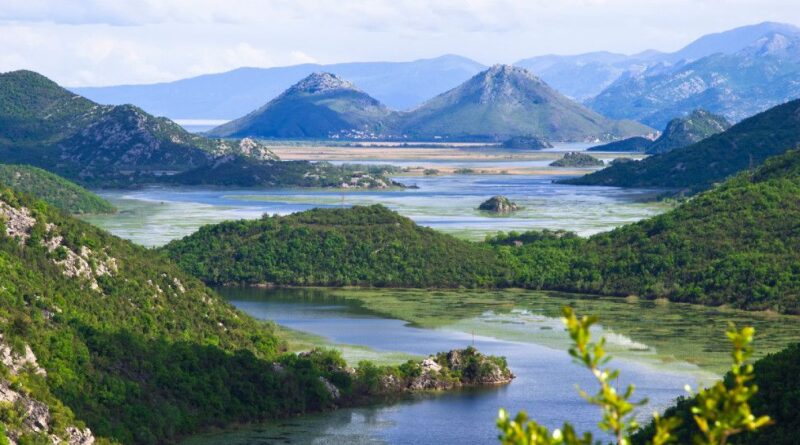 Scenic valley river with green hills, small islands, and distant blue mountains under a partly cloudy sky.