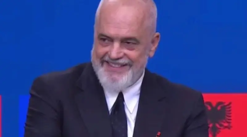 Smiling bald man with gray beard in a dark suit against a blue background.