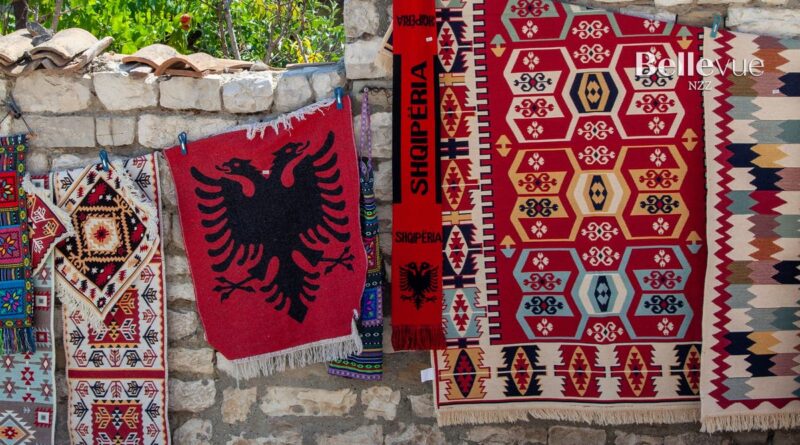 Row of colorful woven rugs hanging on a stone wall, including a red rug with a black double-headed eagle motif.