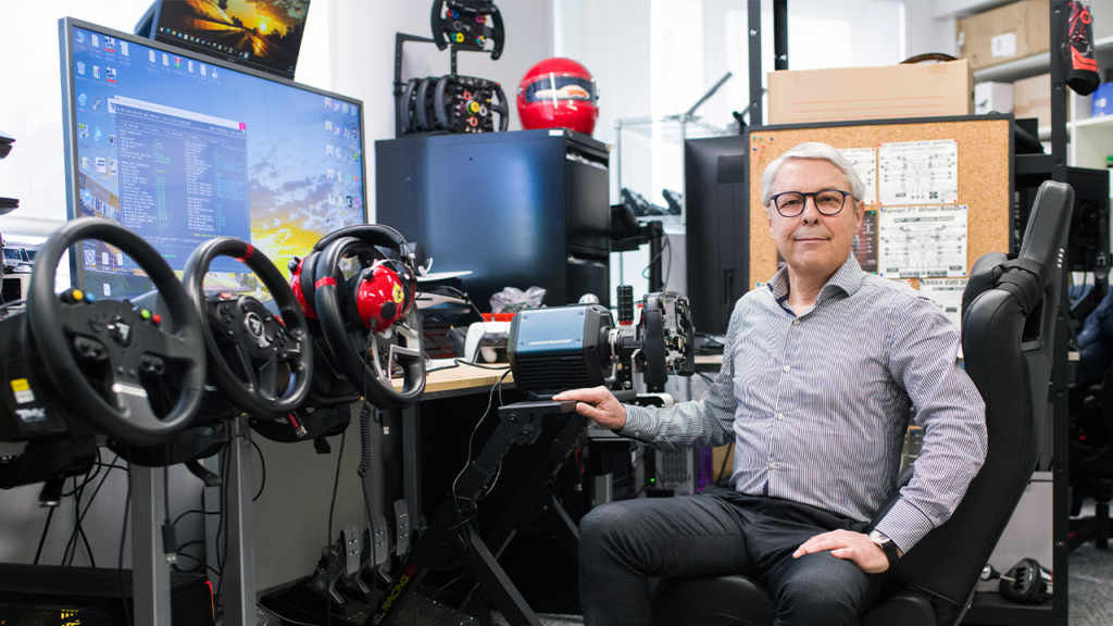 Man with glasses sits at a driving simulator rig with multiple steering wheels and a large monitor in a tech workshop setting, hand on the wheel.