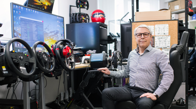 Man with glasses sits at a driving simulator rig with multiple steering wheels and a large monitor in a tech workshop setting, hand on the wheel.