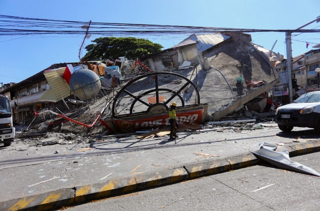 Person in safety gear stands among collapsed buildings and rubble on a city street after a disaster.