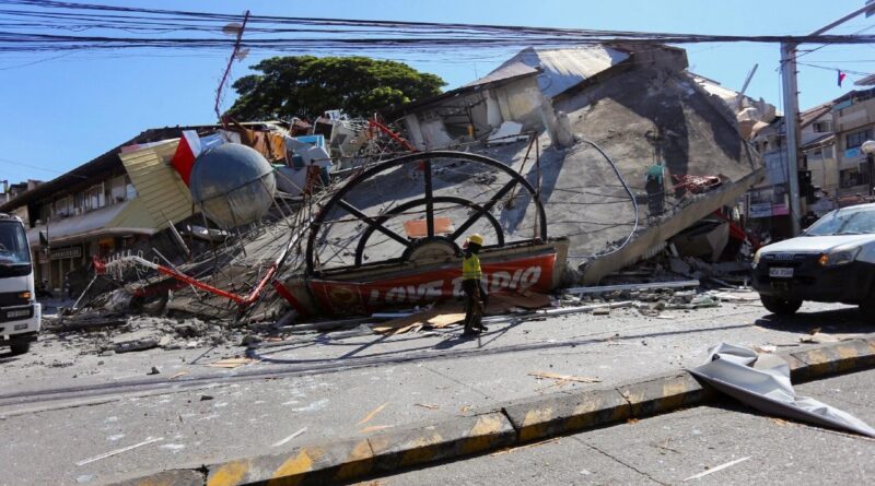 Person in safety gear stands among collapsed buildings and rubble on a city street after a disaster.