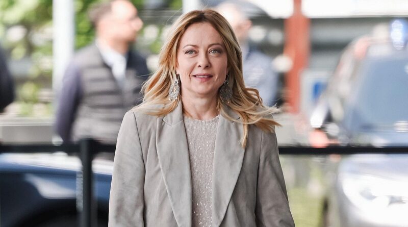 Professional woman walking outdoors in a gray blazer and light sweater, with blurred city background and onlookers behind her.