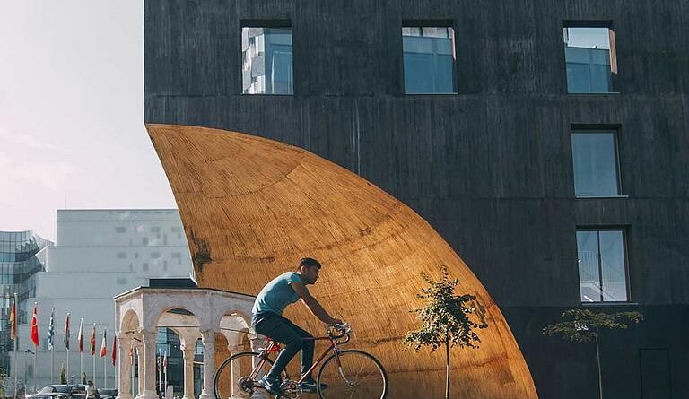 Man riding a bicycle past a large curved wooden arch attached to a dark modern building in an urban plaza. (Informative)