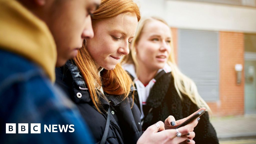 Three young adults outside a brick building looking at a smartphone held by one person, BBC News watermark visible in the corner.