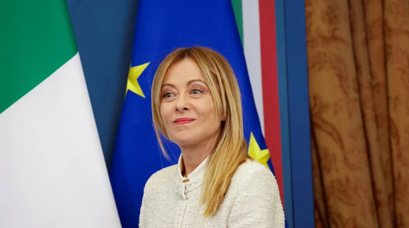 Blonde woman in a light cardigan smiles softly, posing in front of Italian and EU flags.