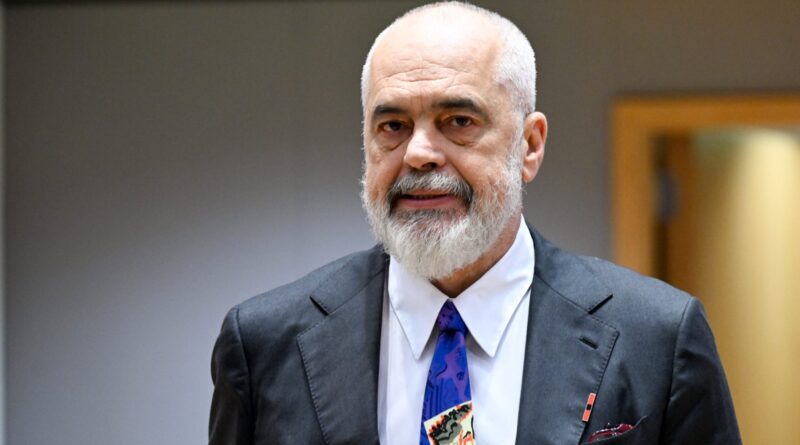 Older man with a gray beard in a dark suit and white shirt, wearing a vibrant patterned tie, looking at the camera.