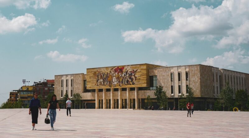 People walk across a wide plaza toward a modern beige building with a large mural on its facade under a blue, partly cloudy sky.