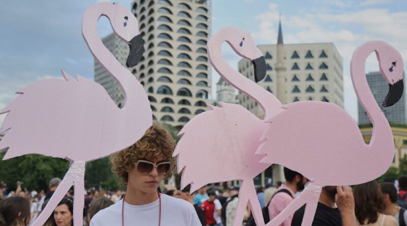 Young person in white shirt and sunglasses stands among oversized pink flamingo cutouts at a city parade.