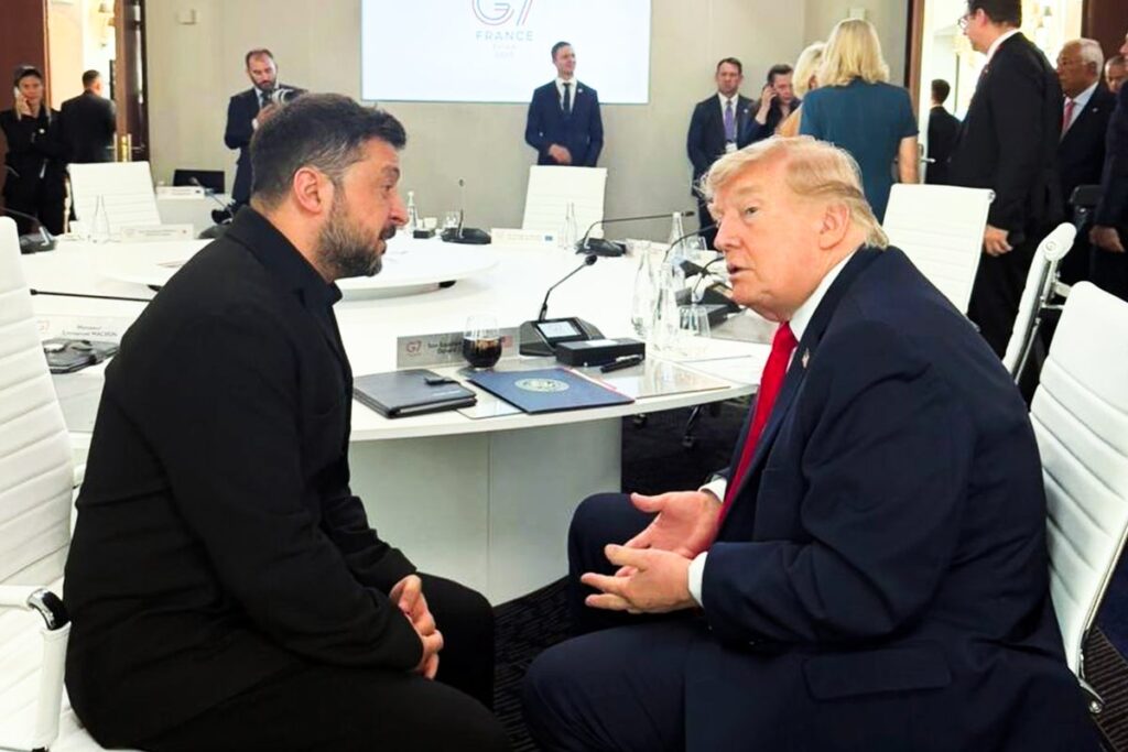 Two men sit opposite each other at a round white conference table, engaged in a serious discussion while others stand in the background in a modern meeting room.