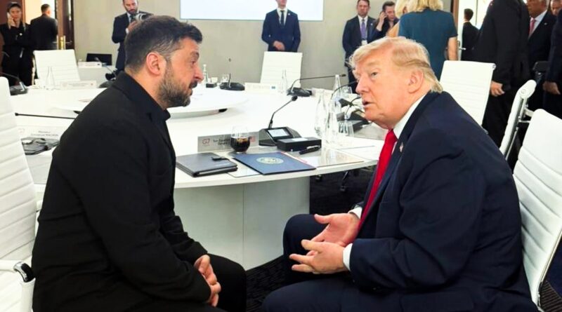 Two men sit opposite each other at a round white conference table, engaged in a serious discussion while others stand in the background in a modern meeting room.