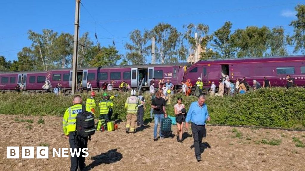 Passengers disembark a halted purple train as police and emergency responders assist on a sunny day.