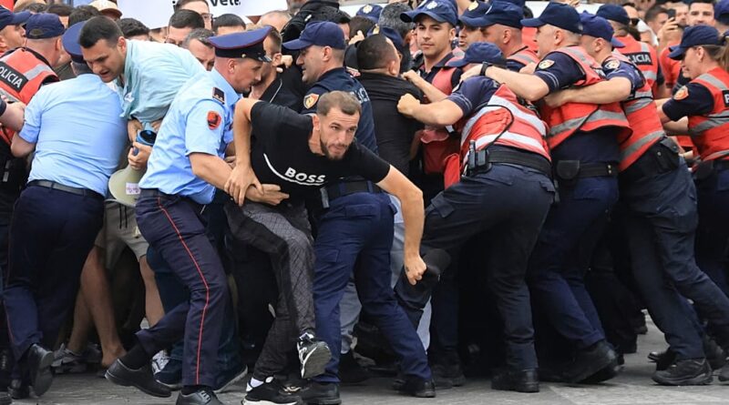 Man in a black shirt labeled 'BOSS' is restrained by police as a chaotic crowd pushes and shoves him.
