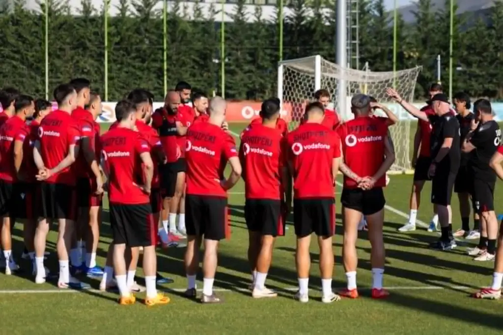 Soccer team in red jerseys standing in a circular huddle on a practice field with a goal in the background.