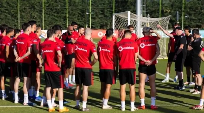 Soccer team in red jerseys standing in a circular huddle on a practice field with a goal in the background.