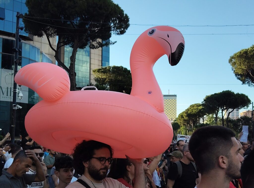 Crowd at a city parade with a giant pink flamingo inflatable towering over spectators behind urban buildings.