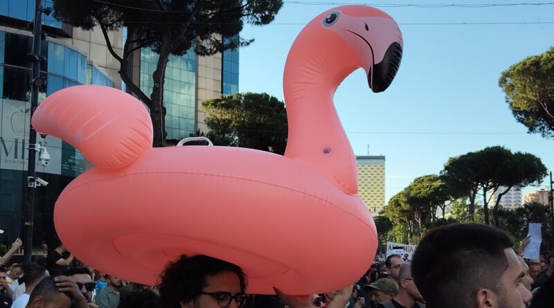 Crowd at a city parade with a giant pink flamingo inflatable towering over spectators behind urban buildings.