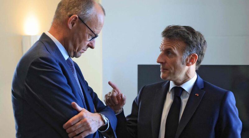 Two men in dark suits talk indoors; one gestures with a raised index finger as the other listens closely.