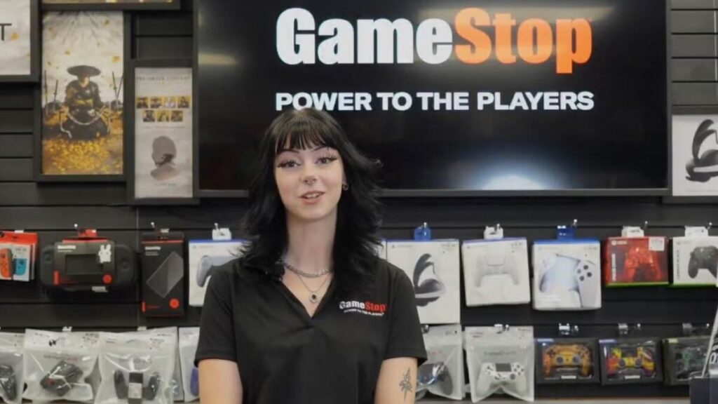 GameStop employee in a black polo stands in front of wall display of gaming accessories, smiling at the camera with a large GameStop sign behind her.
