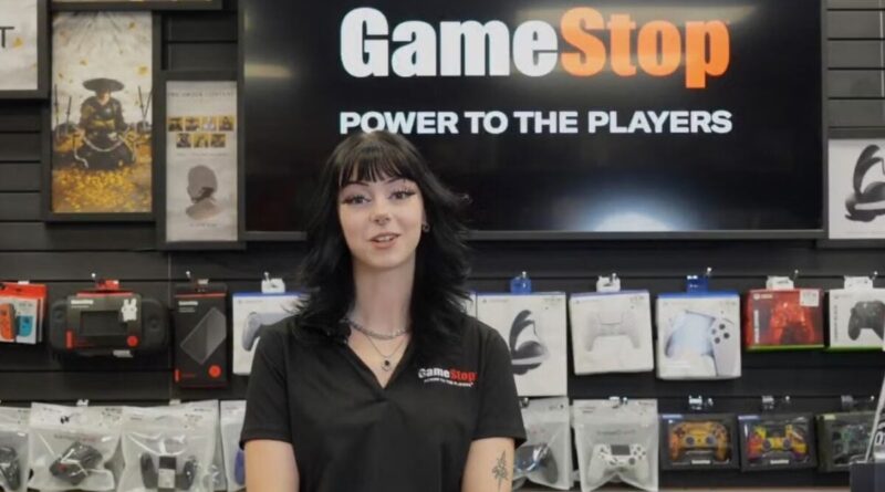 GameStop employee in a black polo stands in front of wall display of gaming accessories, smiling at the camera with a large GameStop sign behind her.