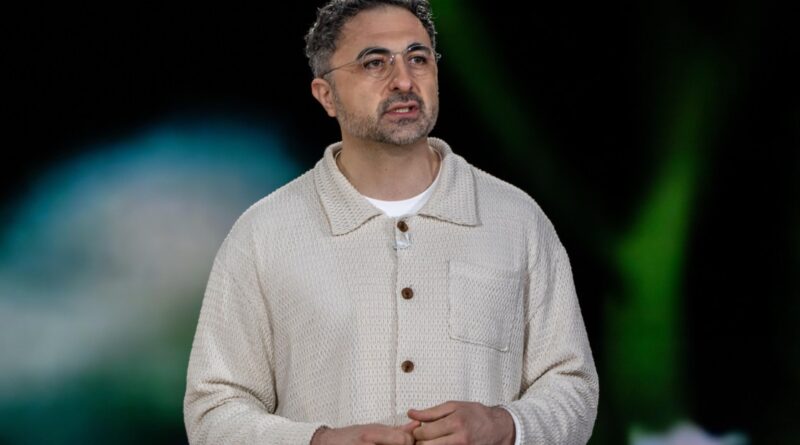 Man wearing a cream knitted cardigan and glasses, speaking on a stage with a blurred background behind him.