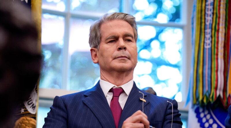 Man in a navy pinstripe suit and maroon tie with a cross lapel pin, eyes closed, at a ceremony with colorful banners in the background.