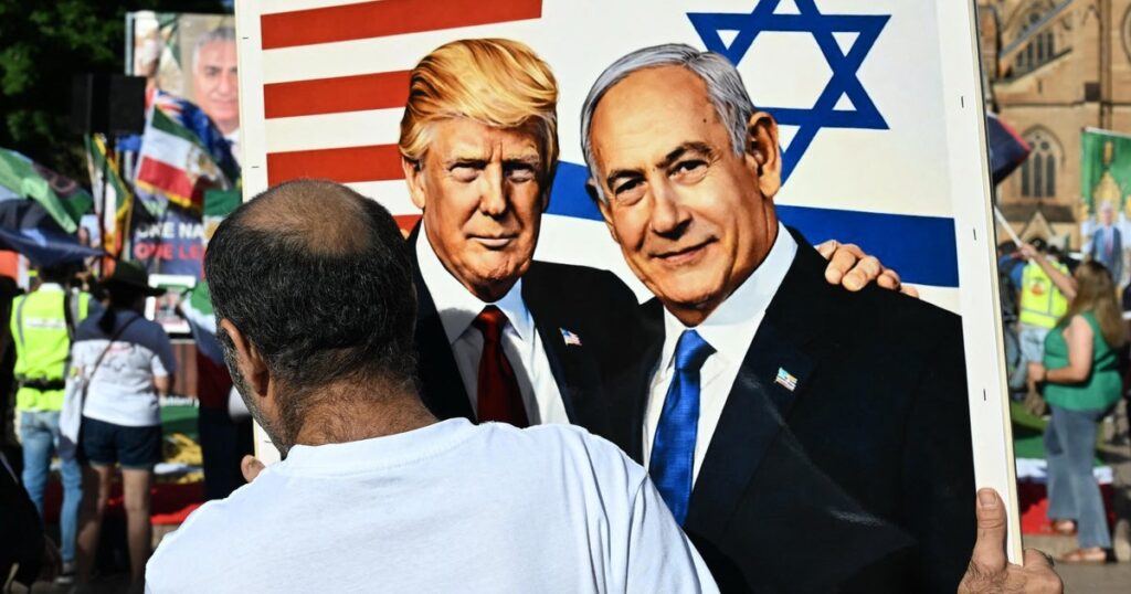 Person viewed from behind holds a large poster of Donald Trump and Benjamin Netanyahu at an outdoor rally, with US and Israeli flags in the background.