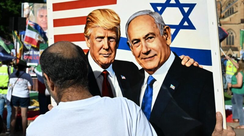 Person viewed from behind holds a large poster of Donald Trump and Benjamin Netanyahu at an outdoor rally, with US and Israeli flags in the background.
