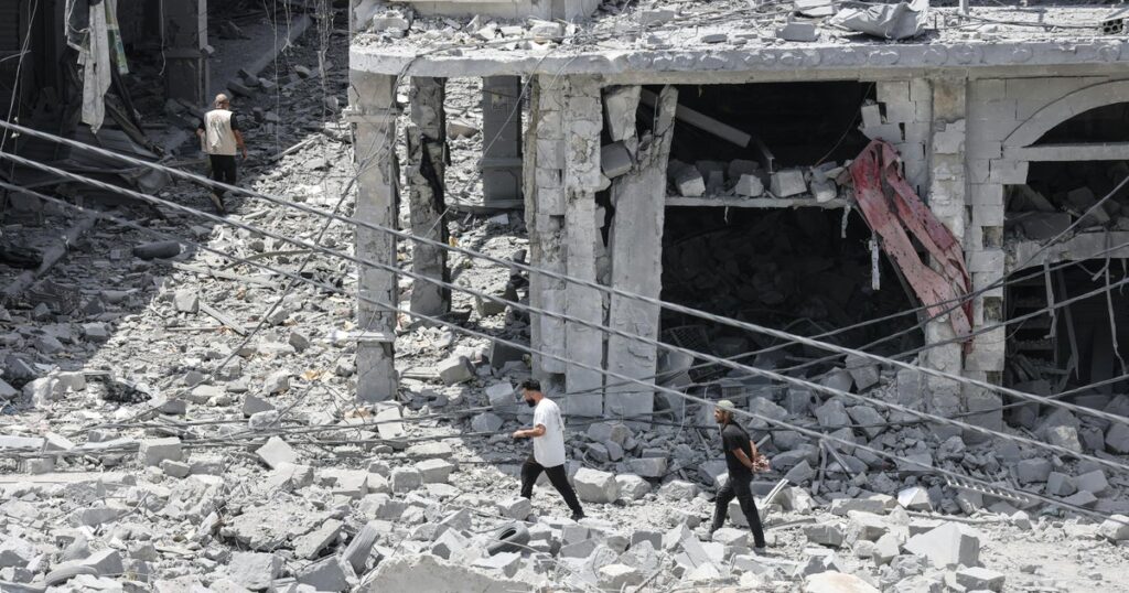 Three men walk through a rubble-strewn street beside a partially collapsed concrete building with exposed pillars and hanging wires in the debris