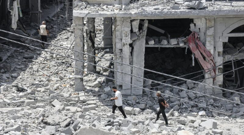 Three men walk through a rubble-strewn street beside a partially collapsed concrete building with exposed pillars and hanging wires in the debris