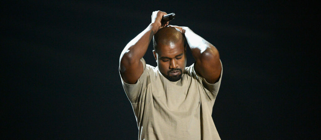 Male performer on stage with arms raised, hands interlaced above head, holding a microphone against a dark background.