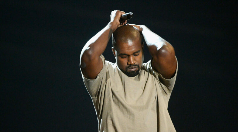 Male performer on stage with arms raised, hands interlaced above head, holding a microphone against a dark background.