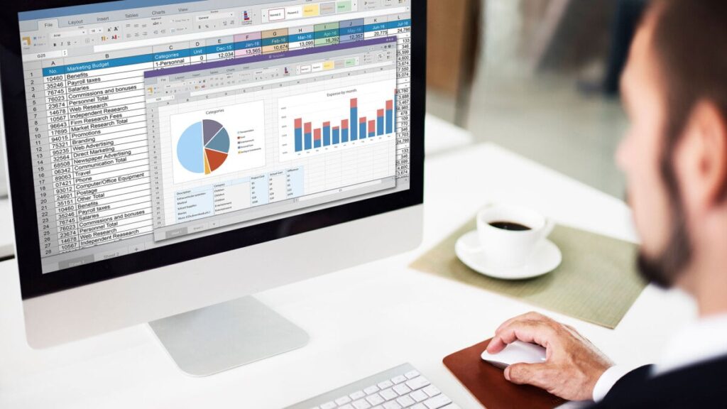 Person working at a desk on an iMac displaying Excel spreadsheets and charts (tables and bar/pie charts).